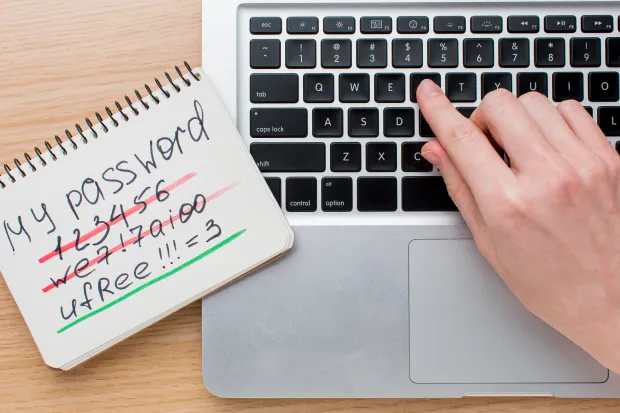 A person typing on a laptop keyboard next to a notepad showing weak and strong password examples, highlighting the importance of secure password creation.